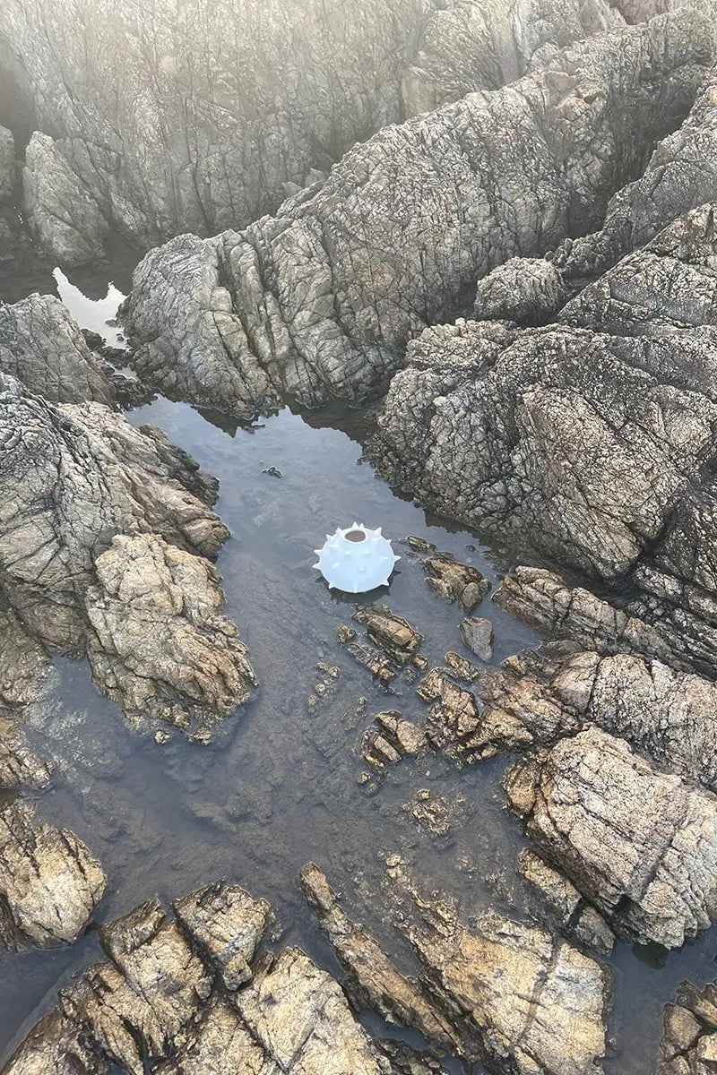 Handmade white round ceramic vase with spikes lying on water amid beach rocks